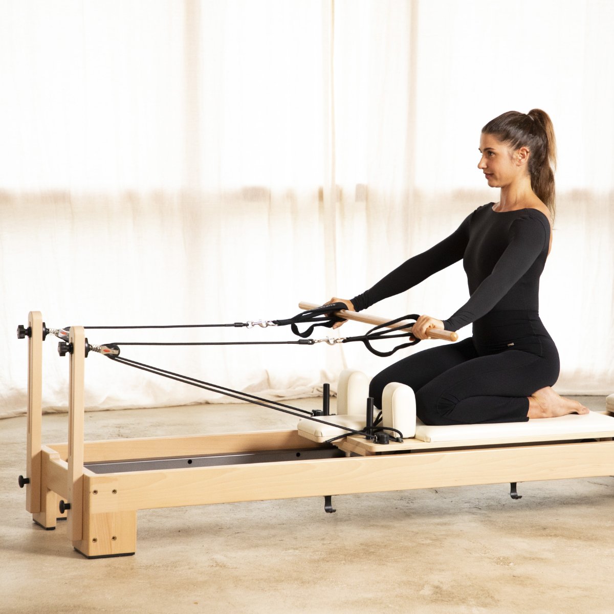 Woman kneeling on the Elina Pilates Lignum Reformer holding the ropes in a bright studio