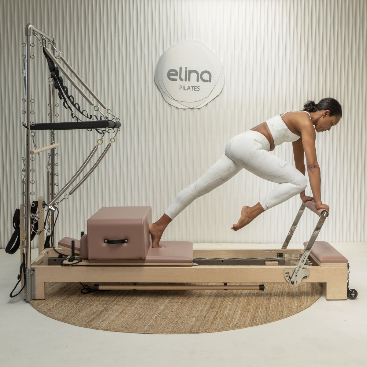 Woman exercising on the Elina Pilates Lignum Reformer with tower and box in front of an Elina Pilates wall