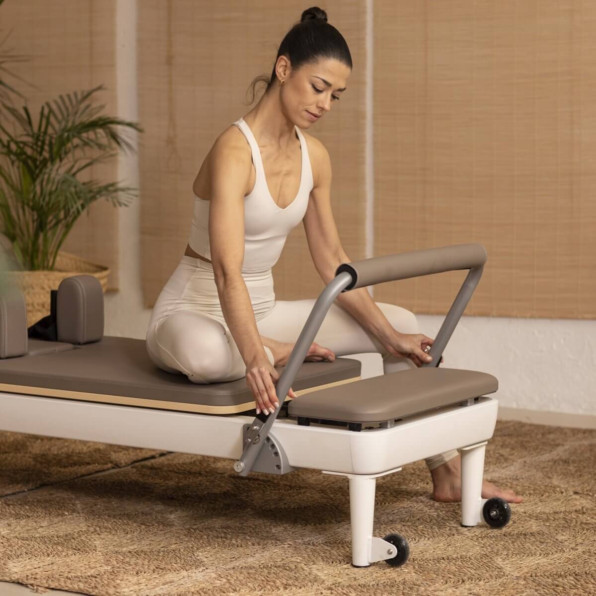Woman sitting on the Elina Pilates Nubium Reformer adjusting the footbar in a studio with plants