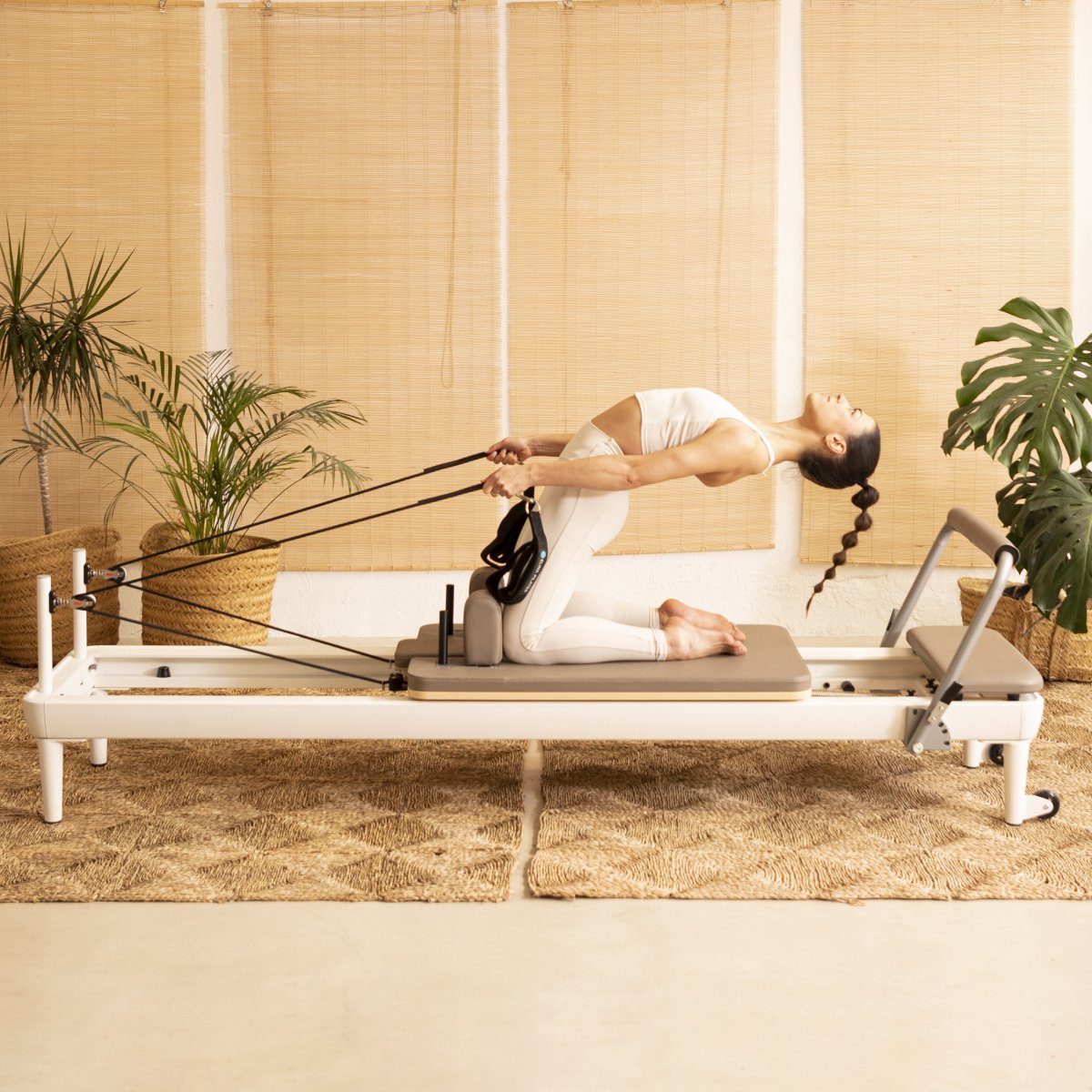Woman exercising on the white-framed Elina Pilates Nubium Reformer in a warm, plant-filled studio